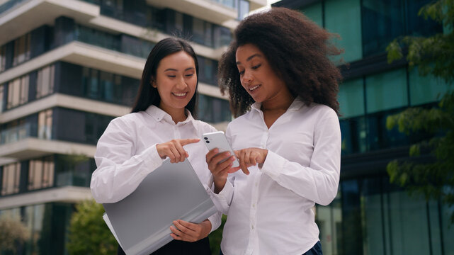 Asian Korean African American women looking mobile phone screen smartphone social media internet girls businesswomen colleagues coworkers fun laughing employees business city cellphone outside females
