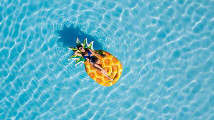 Top view of woman on a big pineapple float in a clear blue pool, shot from drone. Concept summer fun, relaxation, tropical vibes and leisure. © wedmoments.stock