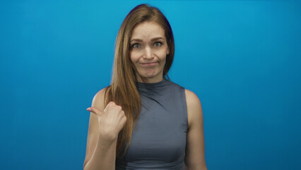 Woman in sleeveless gray top frowning with dramatic thumb pointing to side in blue studio setting;...
