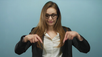 Woman pointing finger downward in blue studio wearing glasses and blazer with white shirt and...