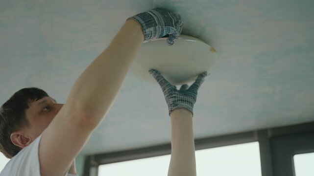 Professional electrician wearing protective gloves installing ceiling light fixture, connecting wiring and mounting electrical component in living room. Concept of electricity maintenance, slow motion - Powered by Adobe