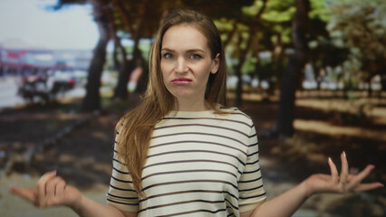 Woman in striped shirt shrugs hands and wrinkles nose in green park beside oak trees under bright...