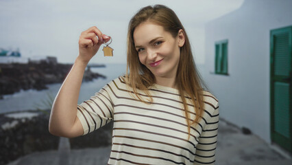 Woman smiling gently holds up tiny wooden house keychain at seaside promenade in bright daylight;...