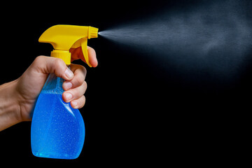Hand holding yellow spray bottle releases fine mist of blue liquid against black background, illustrating cleaning or disinfecting action
