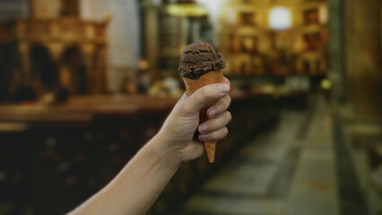 Hand holding chocolate ice cream cone indoors at church setting, symbolizing juxtaposition of...