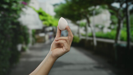Man holding makeup sponge in a city street with blurred greenery background, highlighting urban beauty and everyday life outdoors.