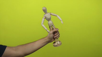 Caucasian man holding a wooden mannequin against a bright yellow wall, emphasizing creativity and design in isolation.
