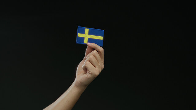 Hand holding a swedish flag patch against a black background, symbolizing swedish nationality and pride, isolated and prominent in the scene, capturing cultural essence and identity.