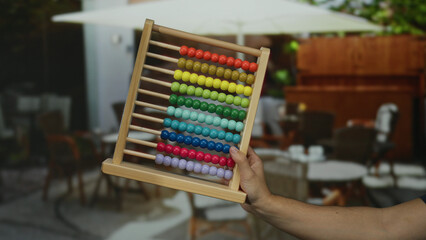 Man holding colorful abacus on outdoor restaurant terrace with blurred background showcasing tables and chairs, evoking a playful educational scene in a city setting.