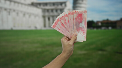 Hand holding pesos in front of pisa tower italy depicting travel and currency exchange by caucasian...