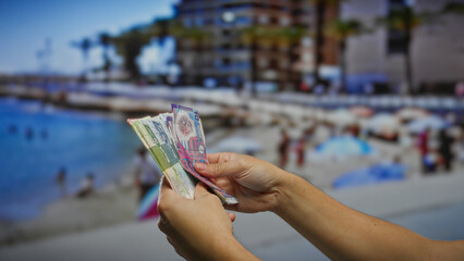 Man holding hong kong banknotes on a sunny seaside street, with blurred beach and buildings in the...