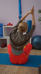 Woman stretching indoors on mat in gym with red pants, focusing on exercise routine amidst gym...