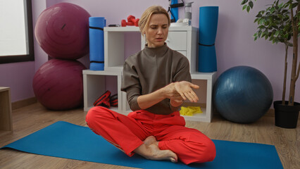 Woman sitting on a yoga mat in a gym holding her wrist, surrounded by fitness equipment, conveys...