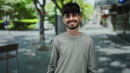 Young hispanic man smiling outdoors in an urban city street with trees in the background, wearing casual clothing.