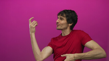 Man smiling while grasping hand with bent fingers and wearing red t shirt in magenta studio;...