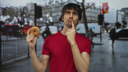 Man in red shirt holding donut with finger on chin on rainy street; thoughtful quiet contemplation.