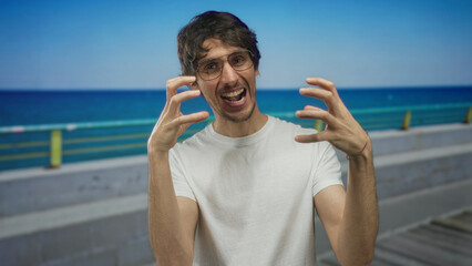 Man wearing glasses and white t shirt grins with claw hands on street by ocean under blue sky;...