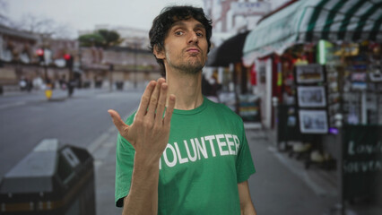 Young man wearing green volunteer shirt beckons with open hand on city street outside souvenir...