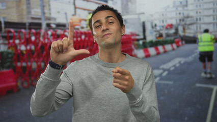 Young hispanic man wearing gray sweater showing loser sign and pointing finger at camera on street;...