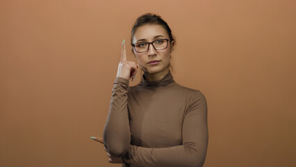 Woman with glasses standing against a brown wall, pointing and thinking, expressing thoughtful and...