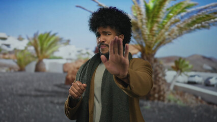 Man gesturing outdoors with palm trees under a clear sky, wearing a jacket and scarf, portraying a...