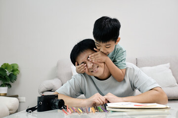 Happy boy covering father eyes while hugging him from behind during playful moment at home