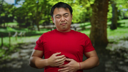Man in red shirt clutches stomach painfully in a sunny park surrounded by lush greenery, highlighting discomfort outdoors.