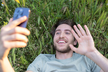 Young man lying down enjoys a video call in a natural setting, showing a cheerful attitude