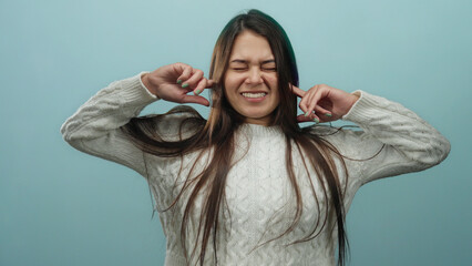 Young hispanic woman in white sweater covers ears with fingers against isolated blue background,...