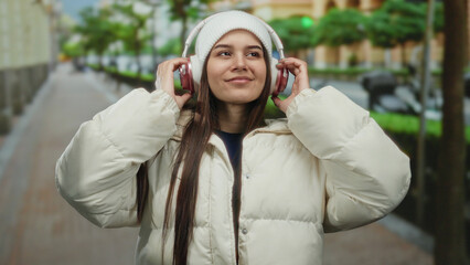 Woman in white jacket with headphones enjoying music on a city street with trees and smiling under...
