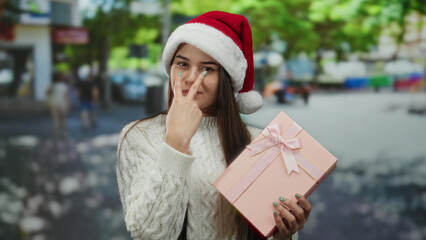 Woman wearing santa hat gestures playful warning holding gift on street in festive outdoor setting,...