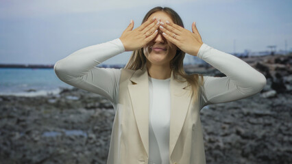 Woman smiling playfully on a rocky seaside in daylight, wearing a beige vest and white outfit, embodying happiness and relaxation near the tranquil sea under a clear sky.
