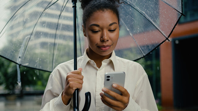 African American woman holding using mobile phone smartphone rain female umbrella texting online internet city typing message technology girl businesswoman business chat cellphone connection outdoor