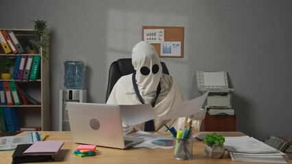 Halloween businessman ghost in white sheet necktie sitting at office desk works with financial...