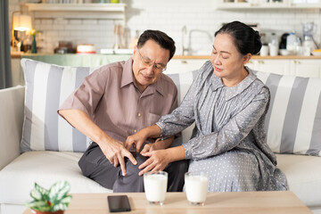 An elderly wife caring for her husband with knee pain on the sofa at home. A tender moment of...