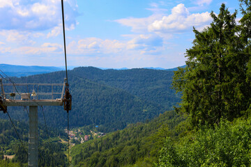 A mountain landscape with dense green forests stretches to the horizon under a blue sky, and a portion of a chairlift in the foreground offers a panoramic view of a picturesque valley with a small set