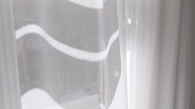 Close-up of white transparent curtain swaying by the wind on a sunny summer day. Modern home interior detail, fresh air coming through the open window. Light fabric moving slowly by the morning breeze