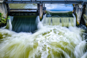 Flowing water cascades over the gates of a hydroelectric dam in a scenic landscape at midday,...
