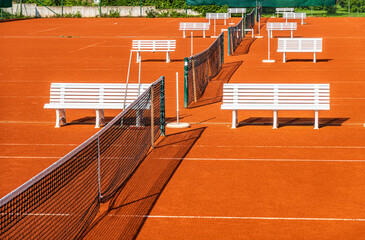 Empty tennis courts with benches on orange clay surface in bright sunlight, creating a serene...