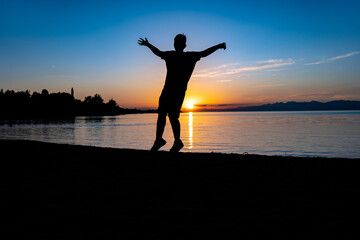 The silhouette of a teenage boy jumping for joy, arms outstretched, on the beach at sunset, with a lake and a bright disc of the setting sun behind him.