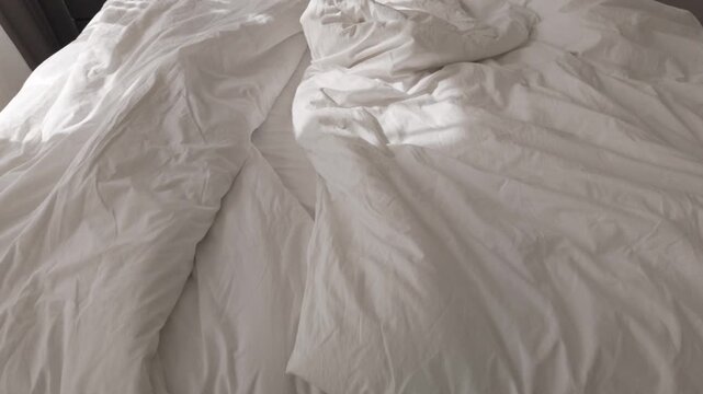 Soft morning sunlight casting shadows on wrinkled white cotton bedsheets. Morning vibes, unmade empty bed. Minimal and cozy home interior. Messy bedroom in a hotel or in an apartment.