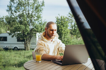 A young man drinks juice and works on a laptop in nature remotely.