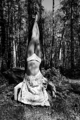 A woman in a floral dress performs a headstand against a tree in a sunlit forest clearing