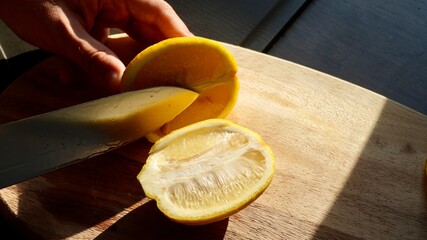 Close-up of hand slicing fresh lemon on wooden board in sunlight, citrus fruit rich in vitamin C, natural ingredient for cooking, drinks, detox, and healthy lifestyle.