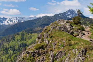 A scenic view of a mountain range with snow-capped peaks, green hills, and a foreground of rocky terrain covered with vegetation in Zakopane - Lesser Poland - Poland