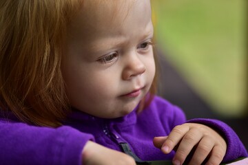 Close-up portrait of thoughtful red-haired toddler girl in purple jacket, focusing intently...