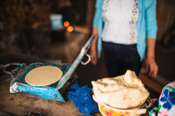 An indigenous Mexican woman wearing a white embroidered blouse and blue sweater, pressing dough into a tortilla press to make tortillas, a traditional Mexican dish.
