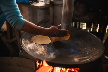 An indigenous Mexican woman wearing a white embroidered blouse and blue sweater, placing flattened dough on the comal to make tortillas, a traditional Mexican dish.
