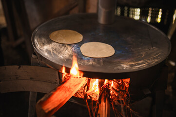 An indigenous Mexican woman wearing a white embroidered blouse and blue sweater, placing flattened dough on the comal to make tortillas, a traditional Mexican dish.

