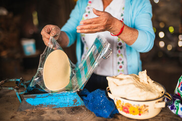 An indigenous Mexican woman wearing a white embroidered blouse and blue sweater, removing the dough from the plastic press to make tortillas, a traditional Mexican dish.

